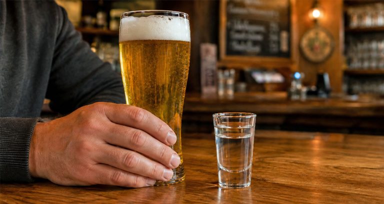 Hand hält ein Bierglas auf der Theke, daneben steht ein Pinnchen Korn.